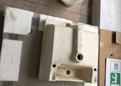 Close-up of bathroom renovation materials, including an old ceramic sink with visible wear, a bucket with a sponge, and packaging materials on a wooden floor.