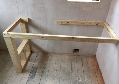 Wooden frame structure built against a plastered wall in a bathroom renovation project, positioned under a window with visible floorboards.