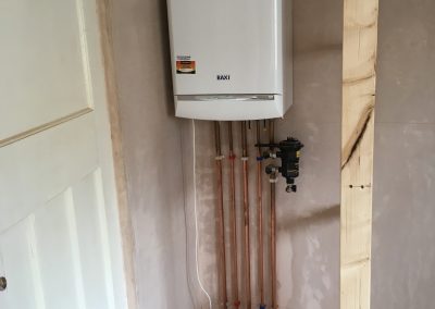Bathroom renovation showing a newly installed Baxi boiler with exposed copper pipes neatly arranged, positioned on a plastered wall, with exposed floorboards below.