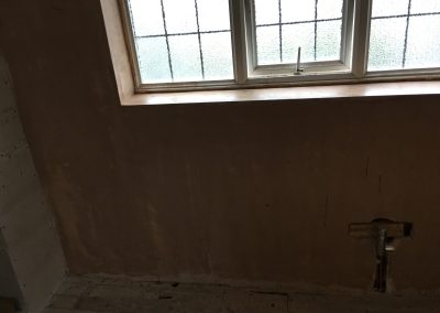 Bathroom renovation progress showing a plastered wall with a frosted window above and exposed plumbing beneath, along with wooden floorboards.