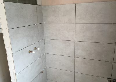 Bathroom wall in progress with light gray tiles partially installed, spacers ensuring alignment, and exposed plumbing fixtures prepared for fitting.