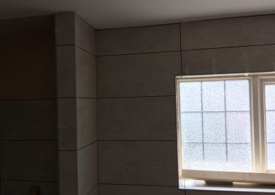 Partially tiled bathroom wall with light gray tiles, showcasing a frosted window and precise corner tiling near the ceiling.