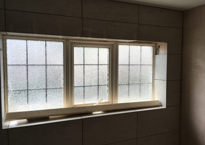 Bathroom wall tiled with light gray tiles, featuring a frosted triple-pane window with partially finished framing.