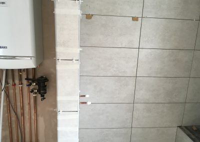 Bathroom renovation featuring light gray tiled walls with visible spacers, a Baxi boiler with neatly aligned copper pipes, and taped-off tiles to secure positioning.